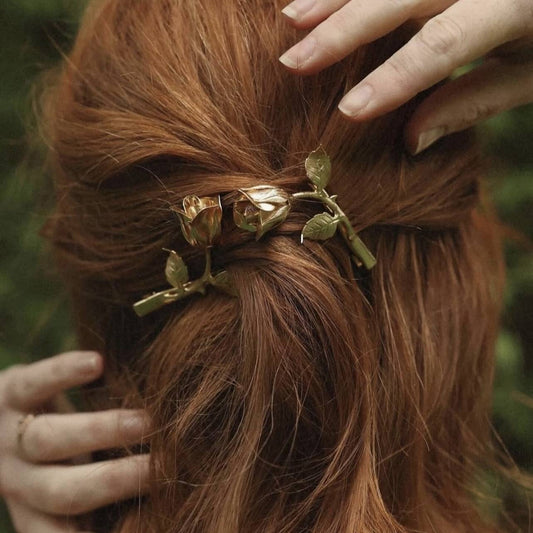 Close-up of a woman’s long hair styled with an elegant gold rose flower hair clip, outdoors against a natural green background.